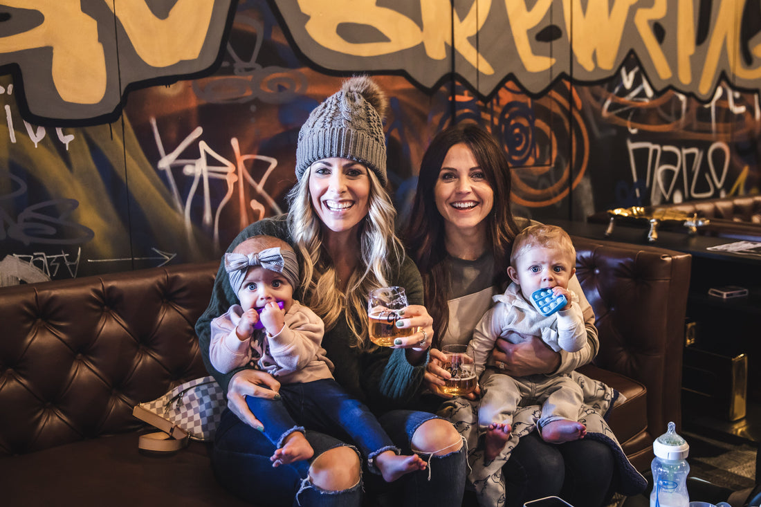 Two women holding NA beer and pregnancy, sitting with children, in a cozy bar setting.